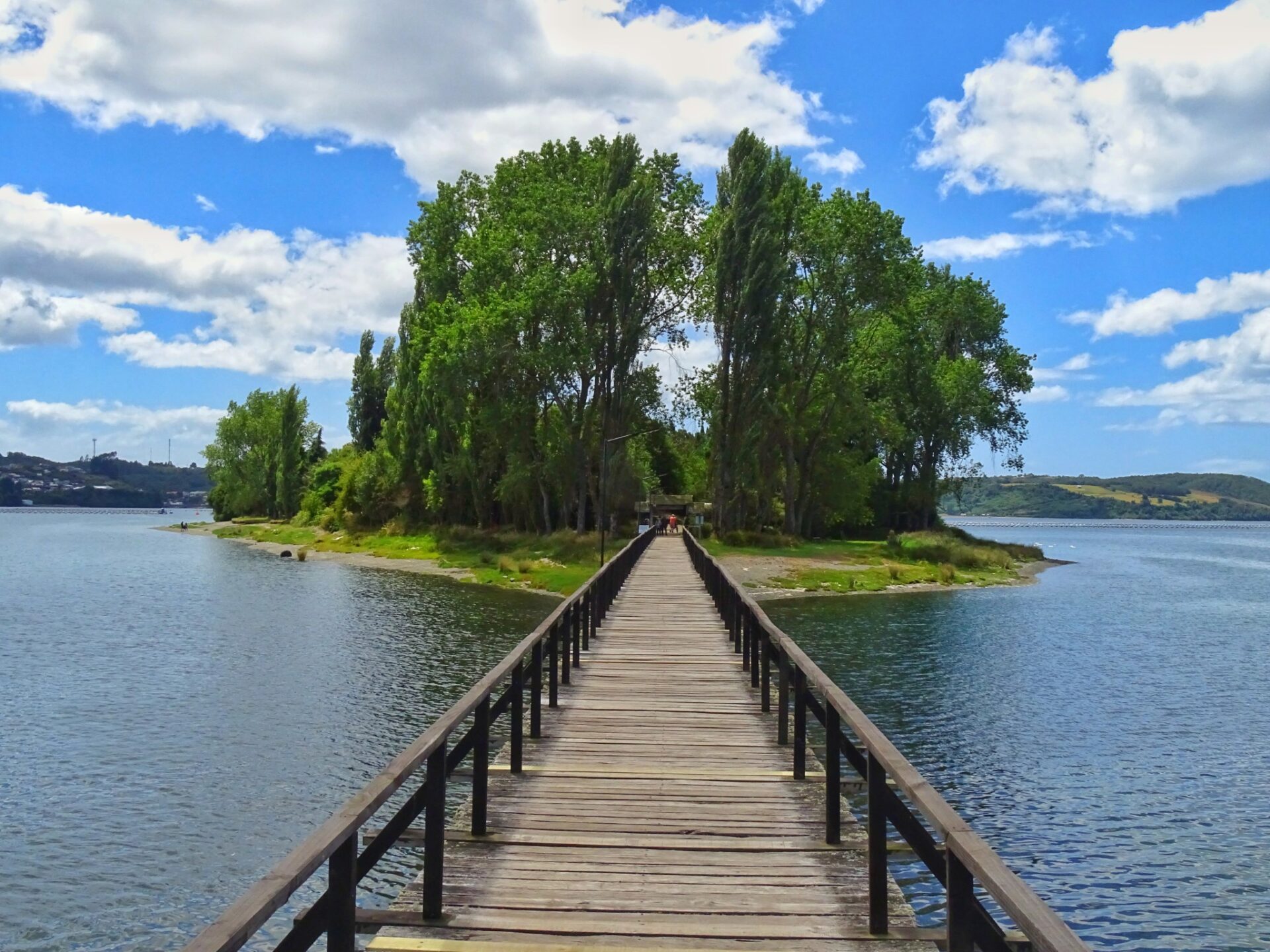 Wooden pedestrian bridge leading to Isla Aucar, surrounded by calm waters and lush trees beneath a bright blue sky with scattered clouds