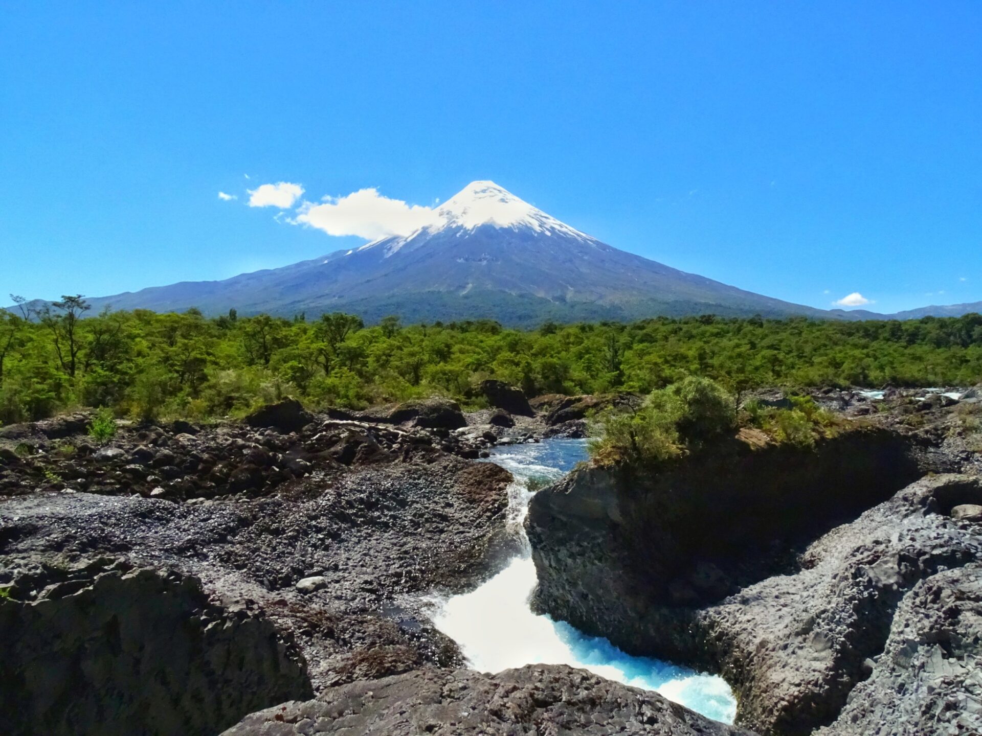 Osorno Volcano rising above a rocky river gorge and forested landscape under clear blue skies.