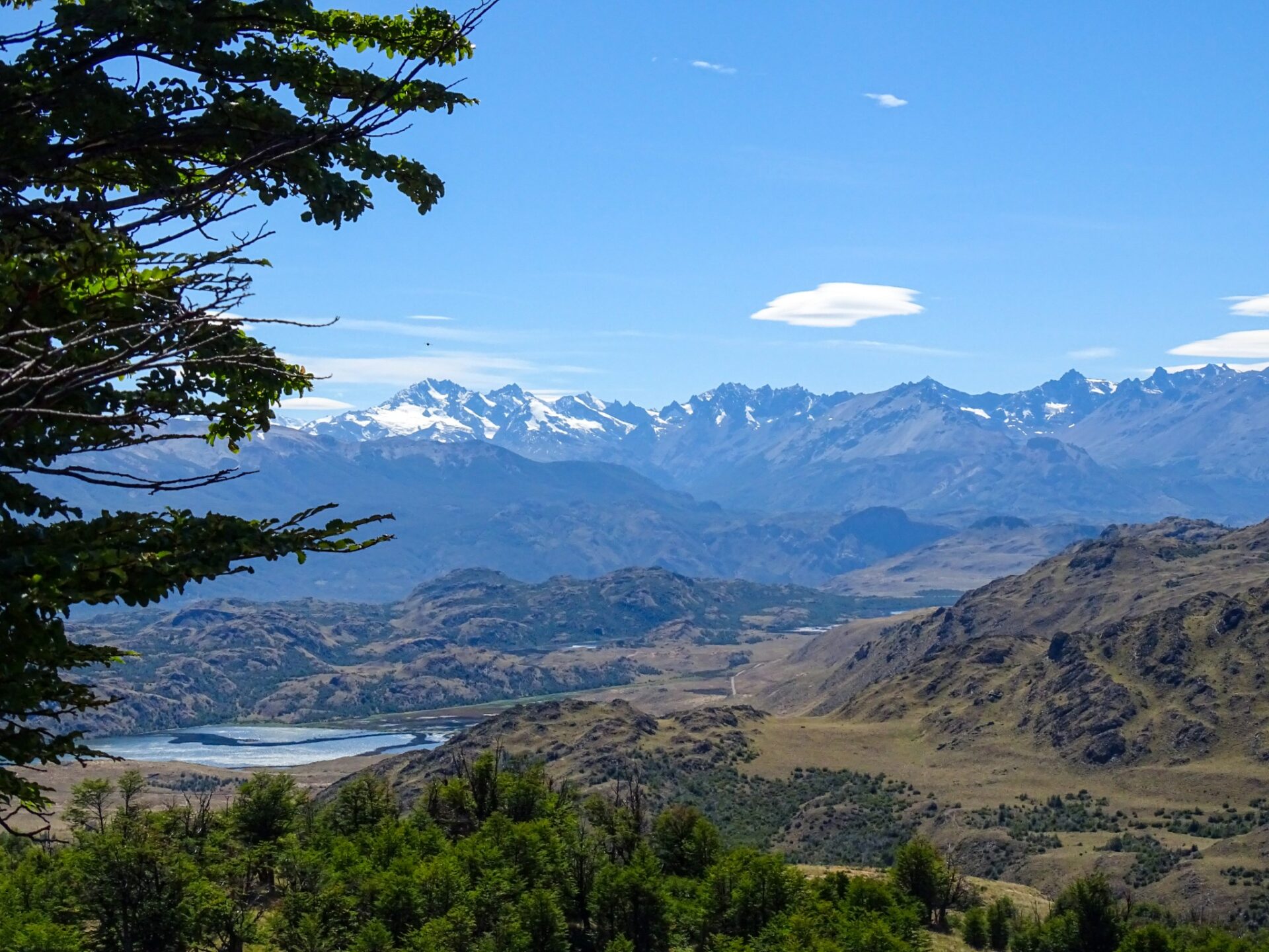 Snow-capped mountain range overlooking rolling hills and a valley landscape beneath a clear blue sky.