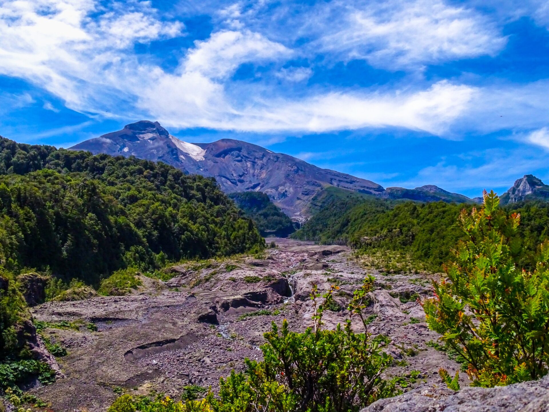Volcanic mountain landscape with forested slopes, rocky terrain, and dramatic clouds overhead.
