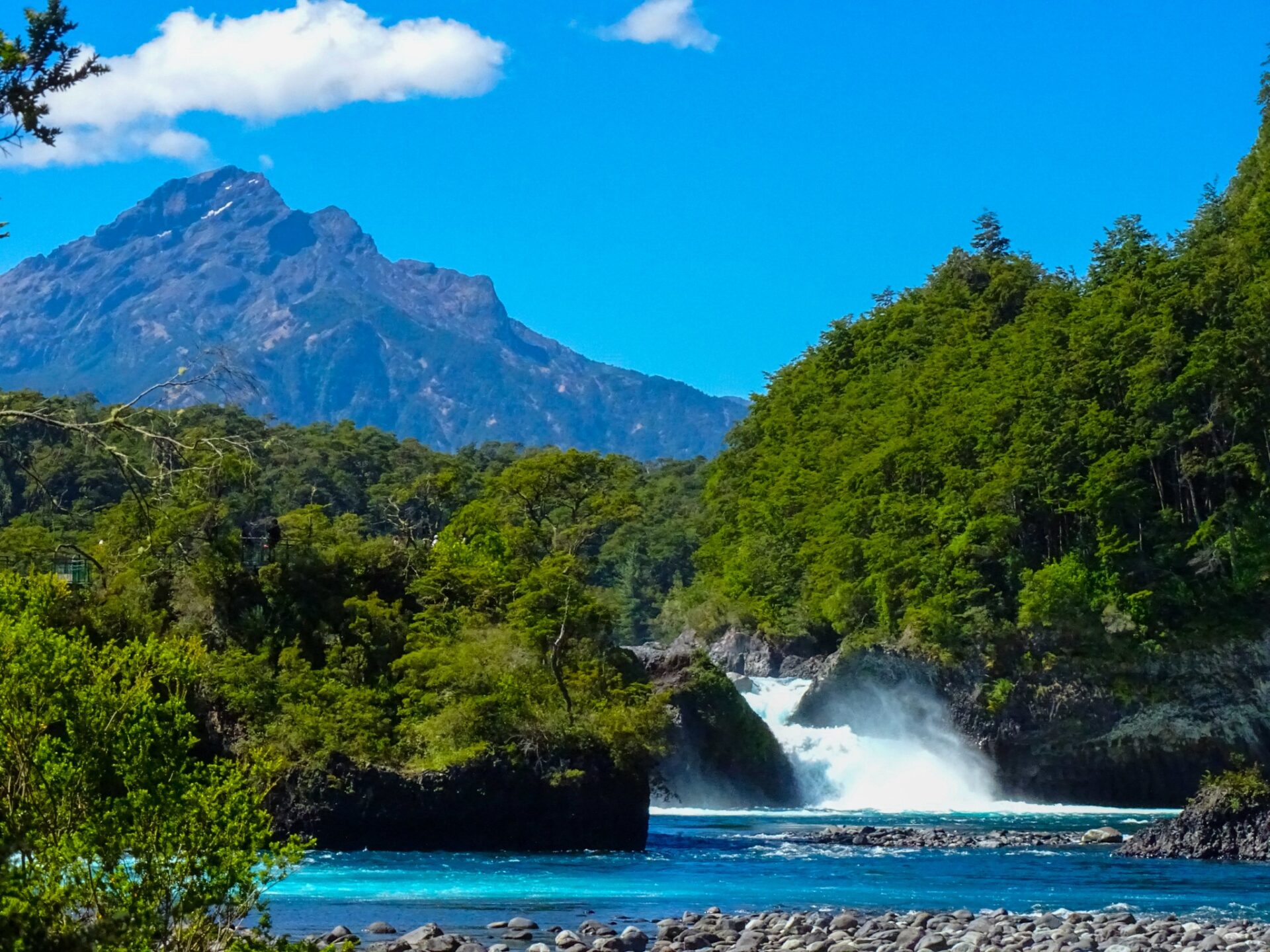 Mountain waterfall flowing through a forested valley beneath a rugged peak and clear blue sky.