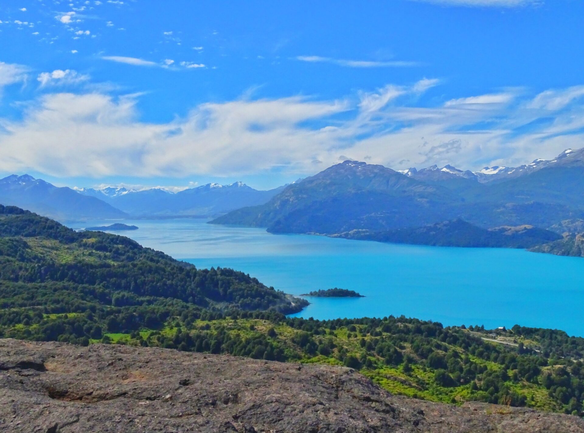 Lago General Carrera surrounded by forested hills and distant mountain ranges beneath a bright sky.